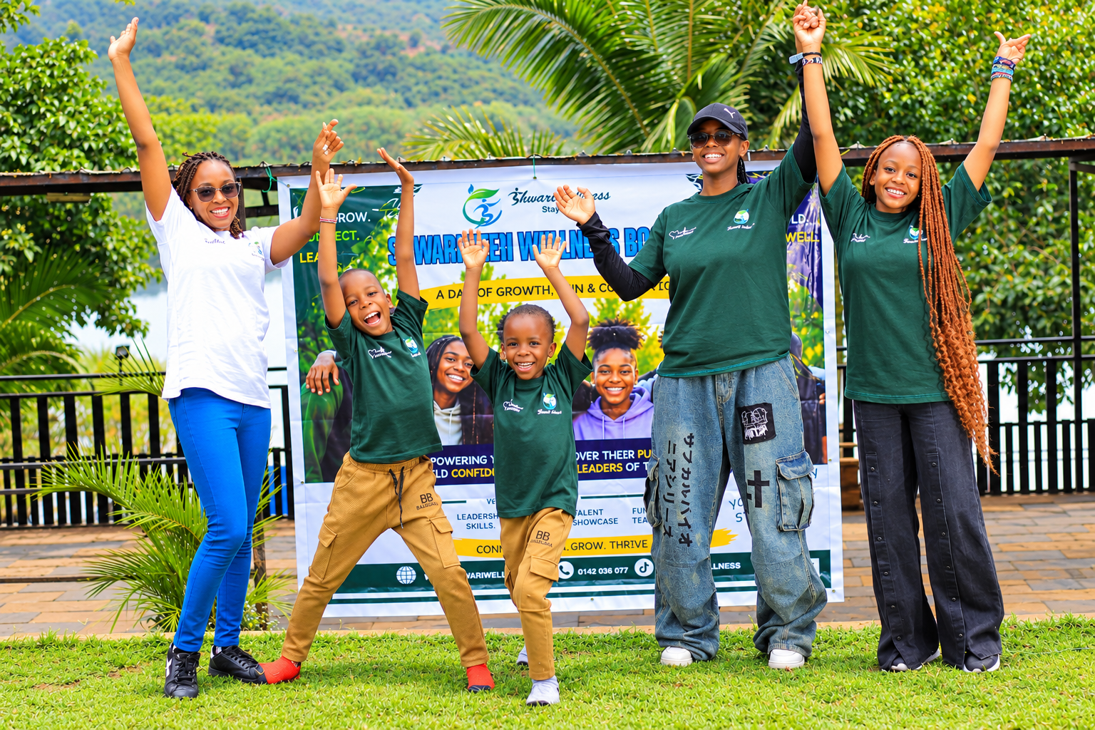 Smiling Shwari Wellness team members and children in Shwari Wellness t-shirts with arms raised in celebration at an outdoor wellness event